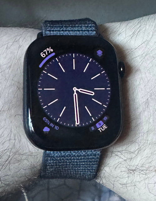 An Apple Watch 10 being worn, with a blue analogue clock on the display, and icons/data shown in the corners