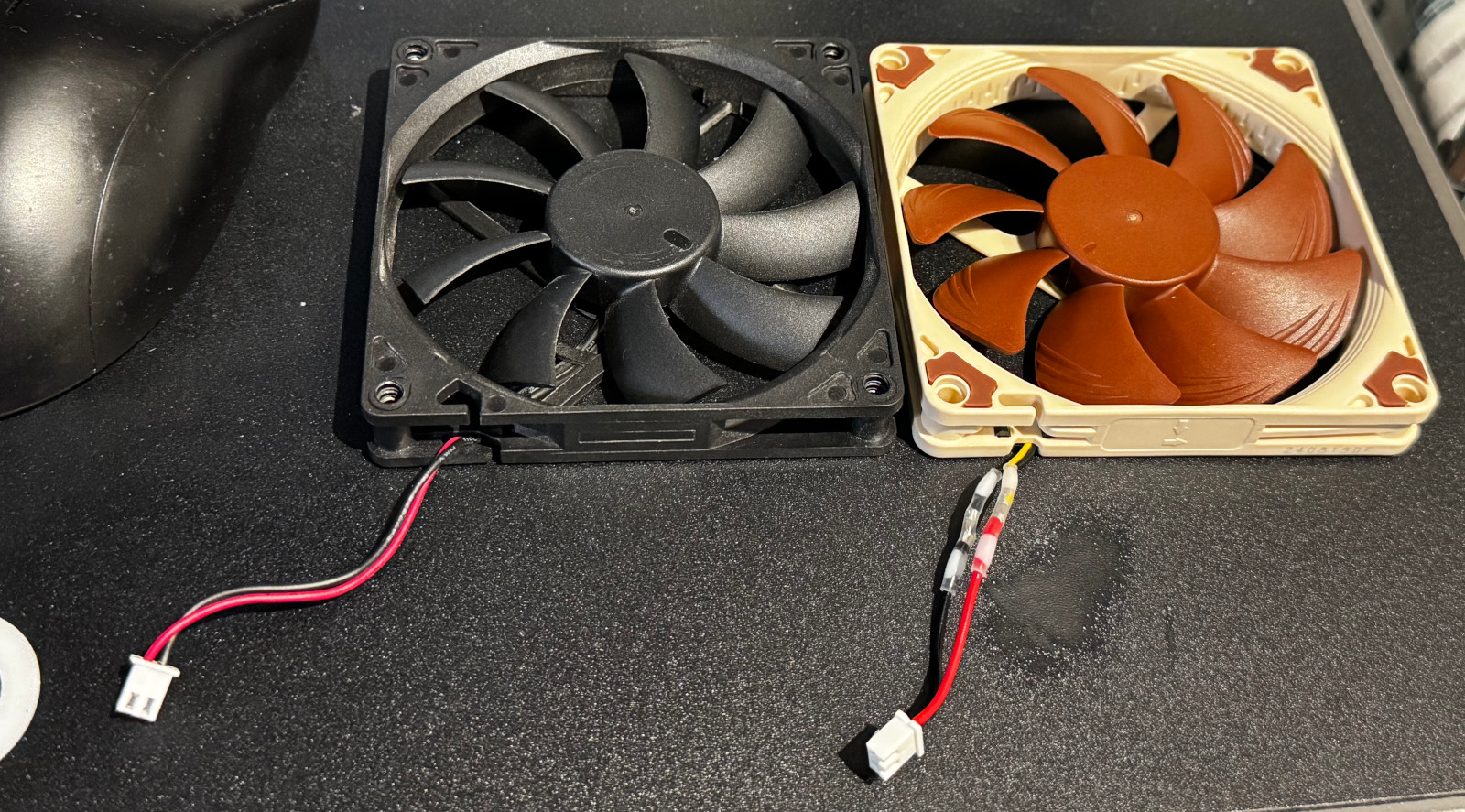 Two fans side by side on a mouse mat: a black fan with a short power connector, and a beige-and-brown fan with transparent solder seals over the joins in its power connector. The mouse mat has a shiny patch near the solder seals.