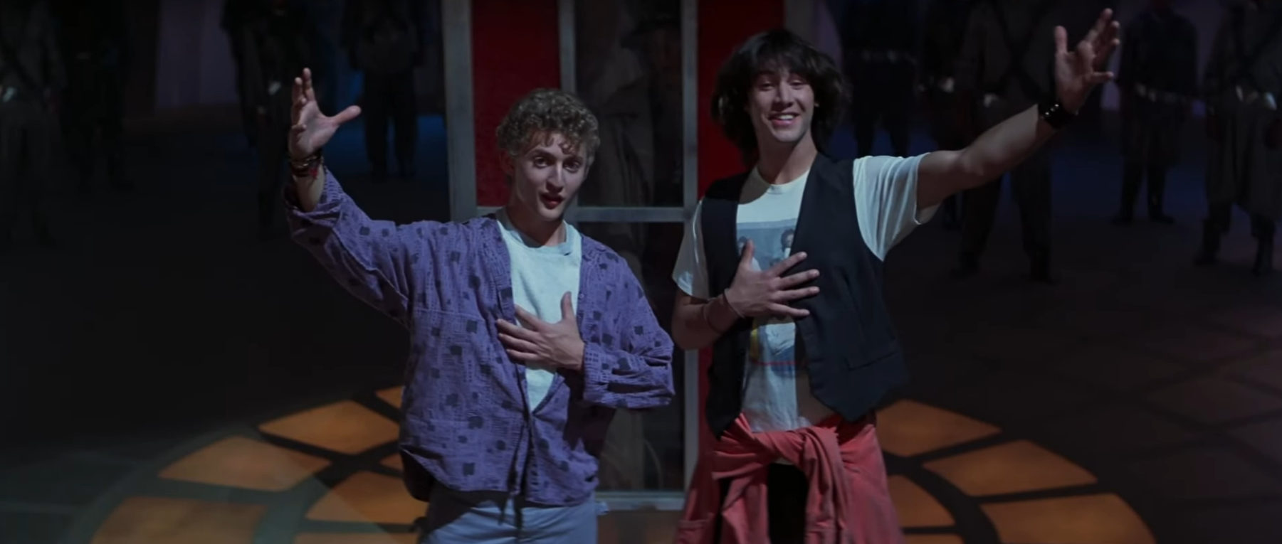 Screengrab from Bill & Ted's Excellent Adventure, with the two stood side by side, one arm stretched out towards the crowd, and the other over their hearts
